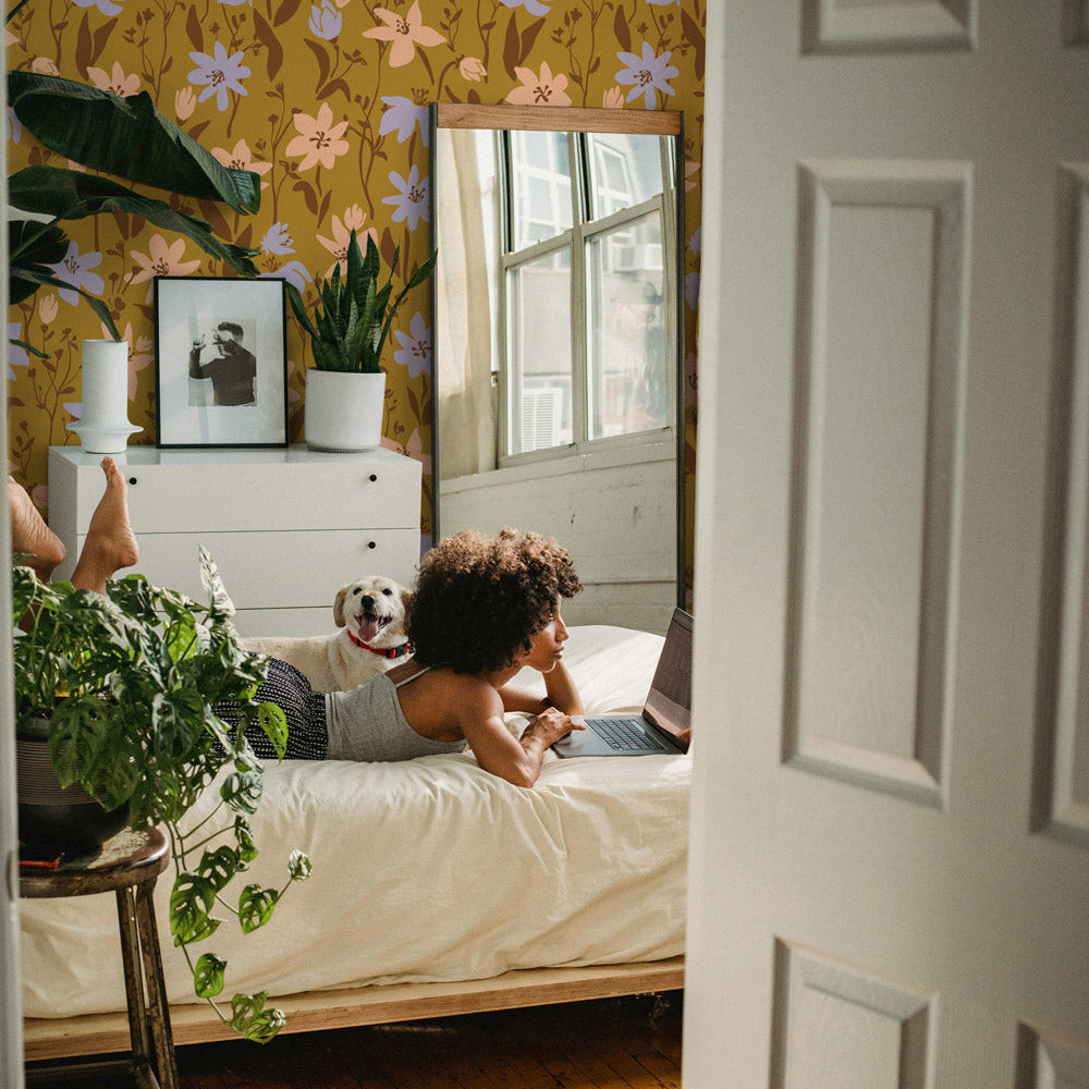 Woman lying on a bed with a dog, surrounded by plants and floral wallpaper.
