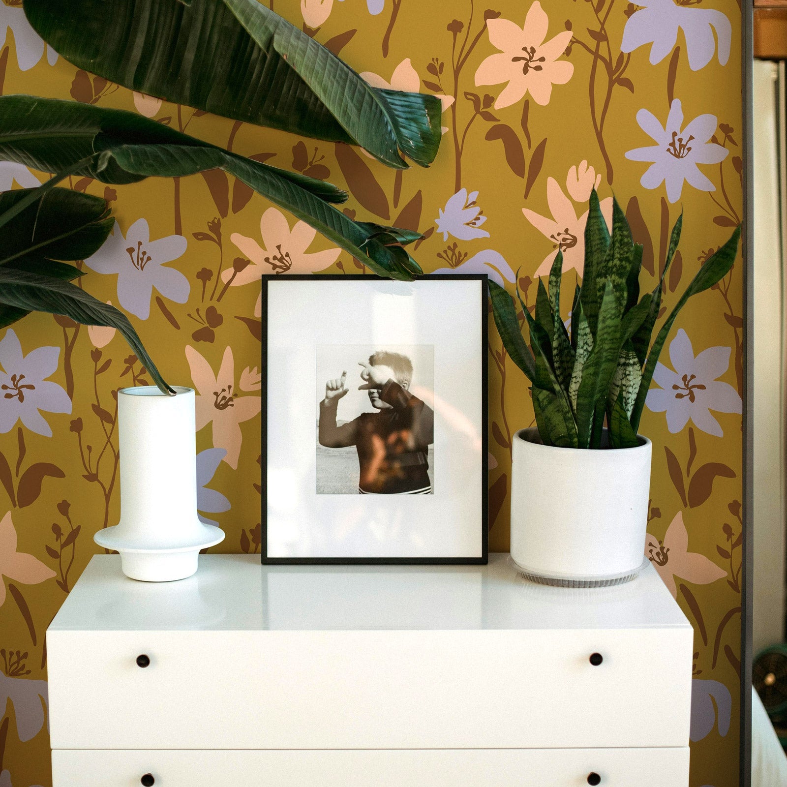 White dresser with a plant, vase, and framed picture against a floral wallpapered wall.